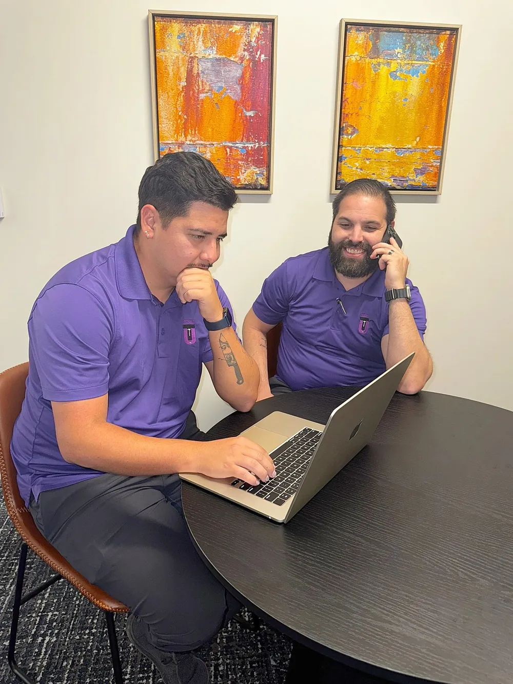 Two members of the Uptown Roofing & Co. team in purple company shirts at a laptop, one of them on the phone