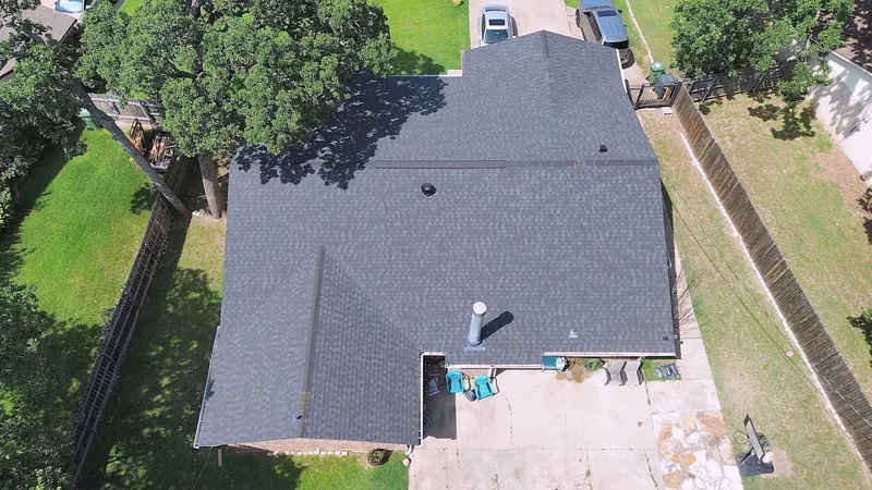 A charcoal shingle roof beside a large oak tree, over a home with a back patio, from the air