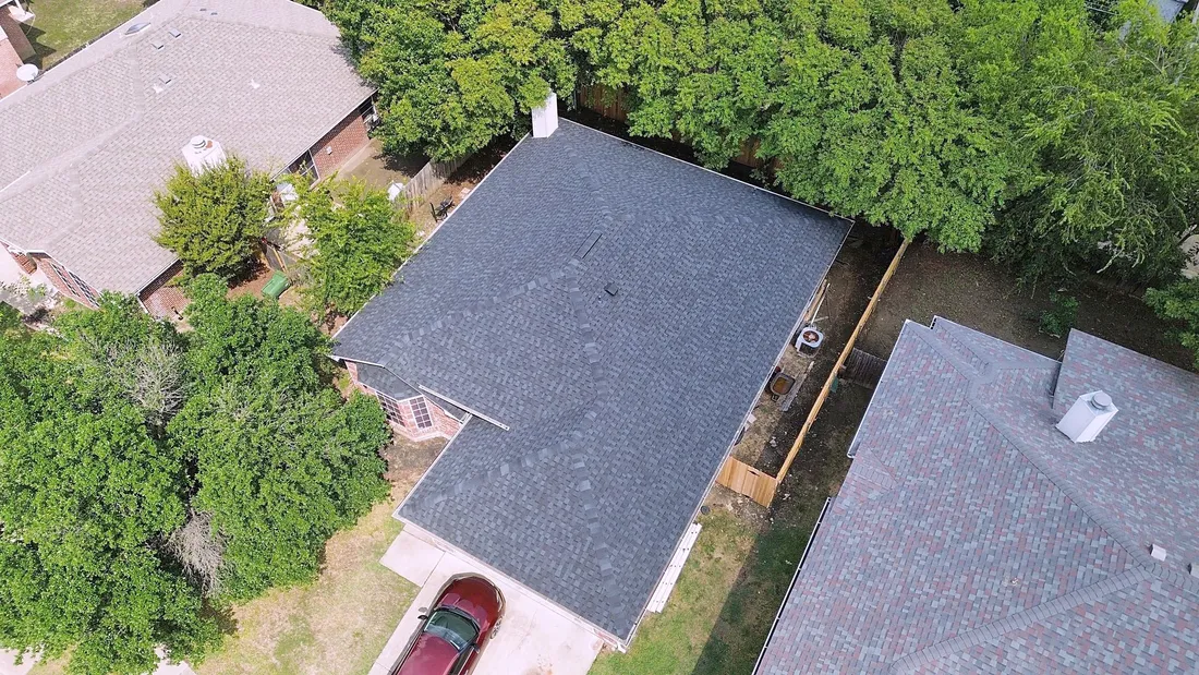 A charcoal gray shingle roof on a brick home backed by tall trees, with a red car in the driveway, from the air