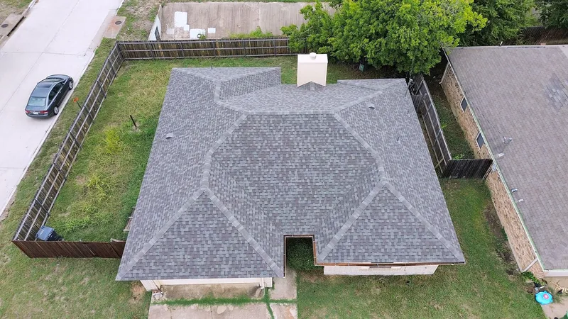 A gray shingle hip roof over a home with a wood privacy fence around the back yard, from the air