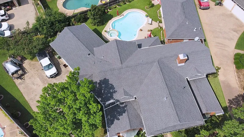A large gray shingle roof with several gables and a chimney, beside a backyard pool, from the air