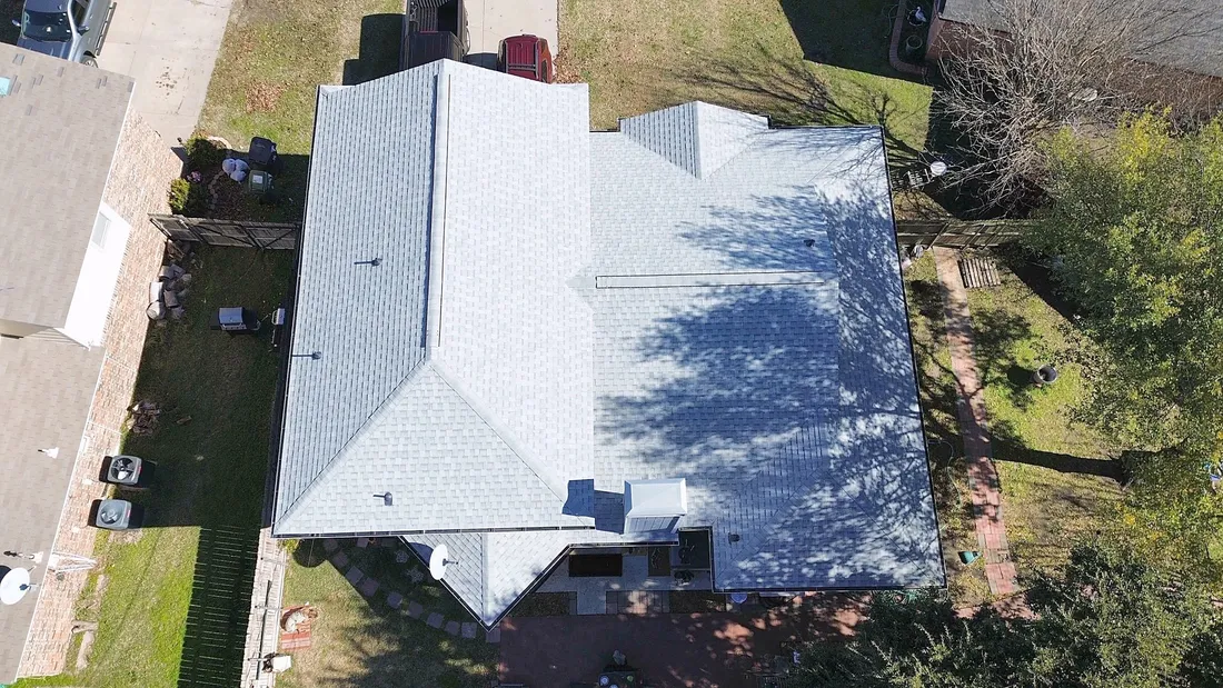A light gray shingle roof partly in the shade of a large tree, from the air
